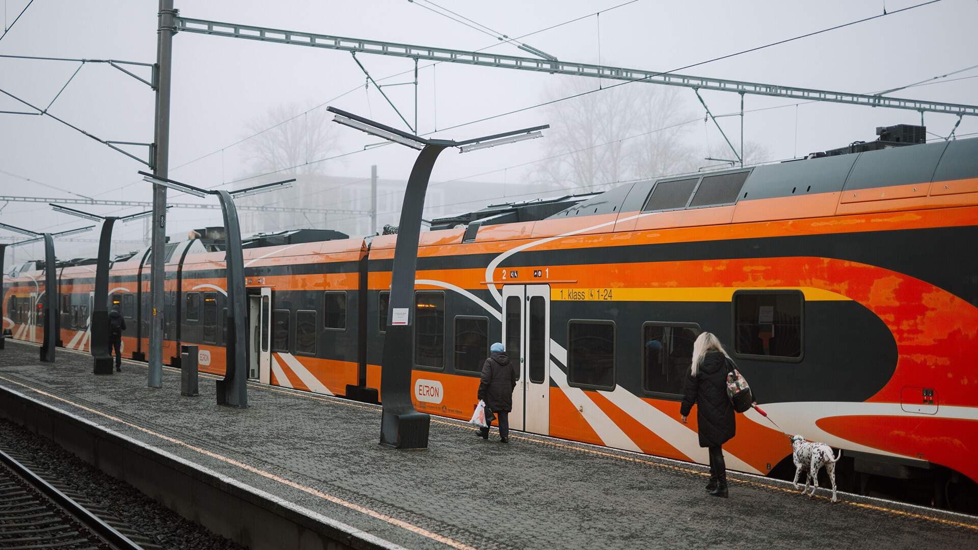 Orange Elron train at a concrete railway platform with overhead wires and tracks on the Tallinn–Tartu line.