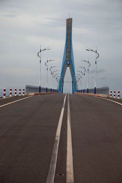 Aerial view of a coastal bridge crossing a bay, with a distinctive blue pylon and empty roadway under an overcast sky.