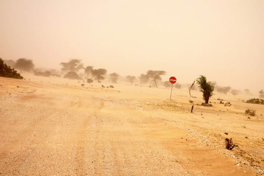 A dusty, unpaved road winds through an arid landscape in Mauritania, featuring a stop sign and sparse vegetation under hazy, sand-filled skies.