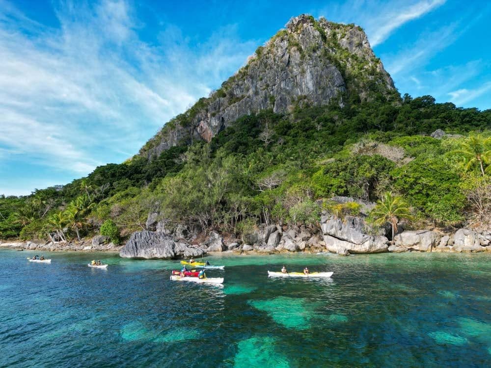 Kayakers paddle in turquoise waters near the lush, rocky coastline of Sawa-i-Lau island off Labasa under a blue sky with scattered clouds.