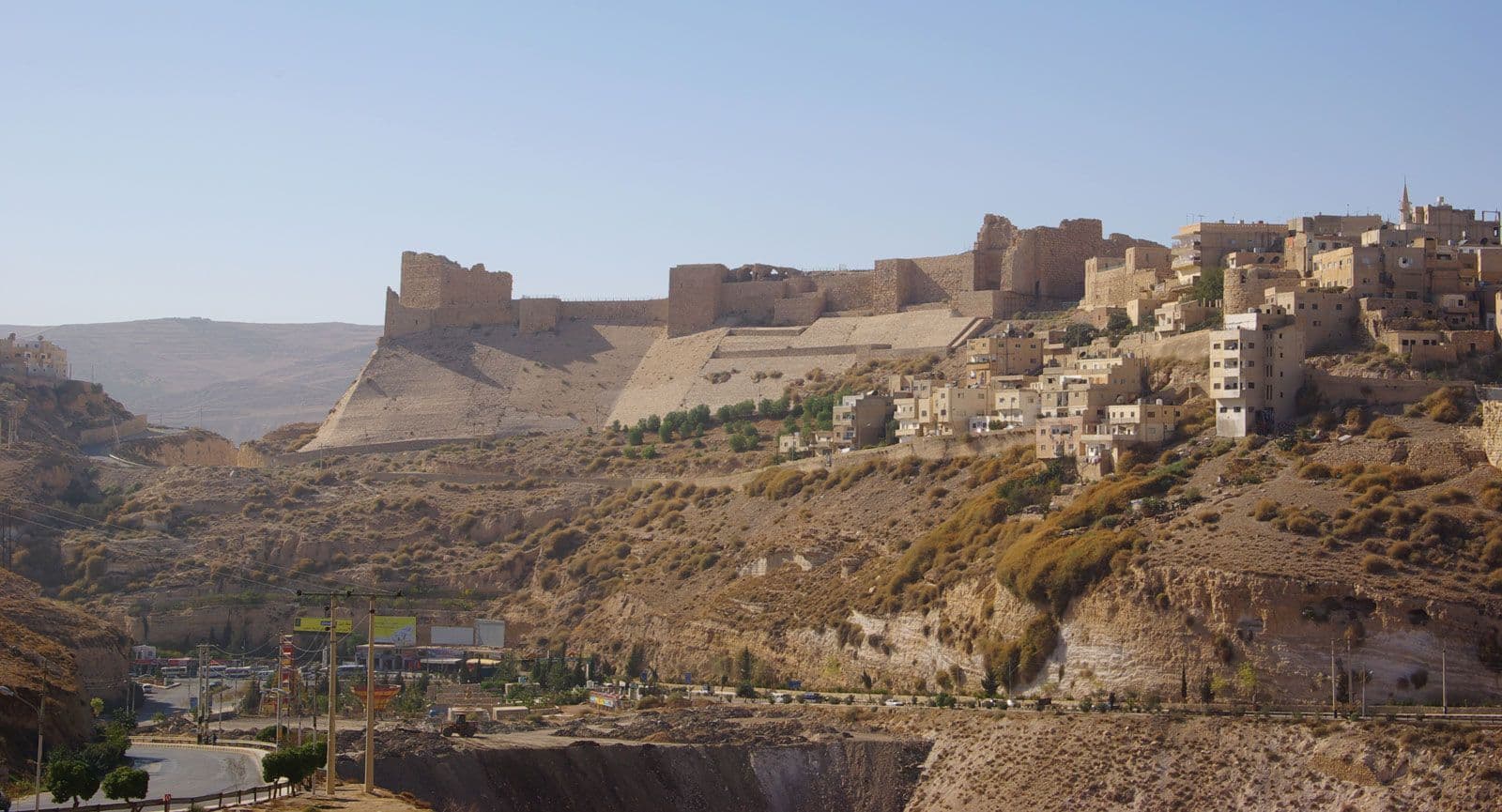 A wide landscape view of the Karak Citadel and surrounding stone village buildings situated on a rugged, arid hillside under a clear blue sky.