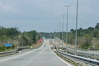 A wide, multi-lane highway stretches into the distance under an overcast sky, flanked by metal guardrails, tall streetlights, and dense green vegetation.