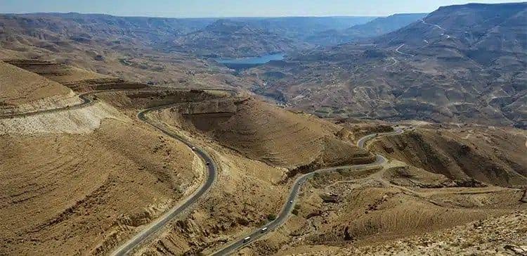 Aerial view of a winding road through arid, mountainous terrain in the Karak region, Jordan, with a distant lake and valley.