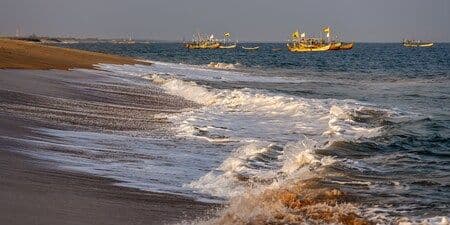 Coastal shoreline near Anloga with breaking waves and several yellow fishing boats anchored offshore under a clear sky.
