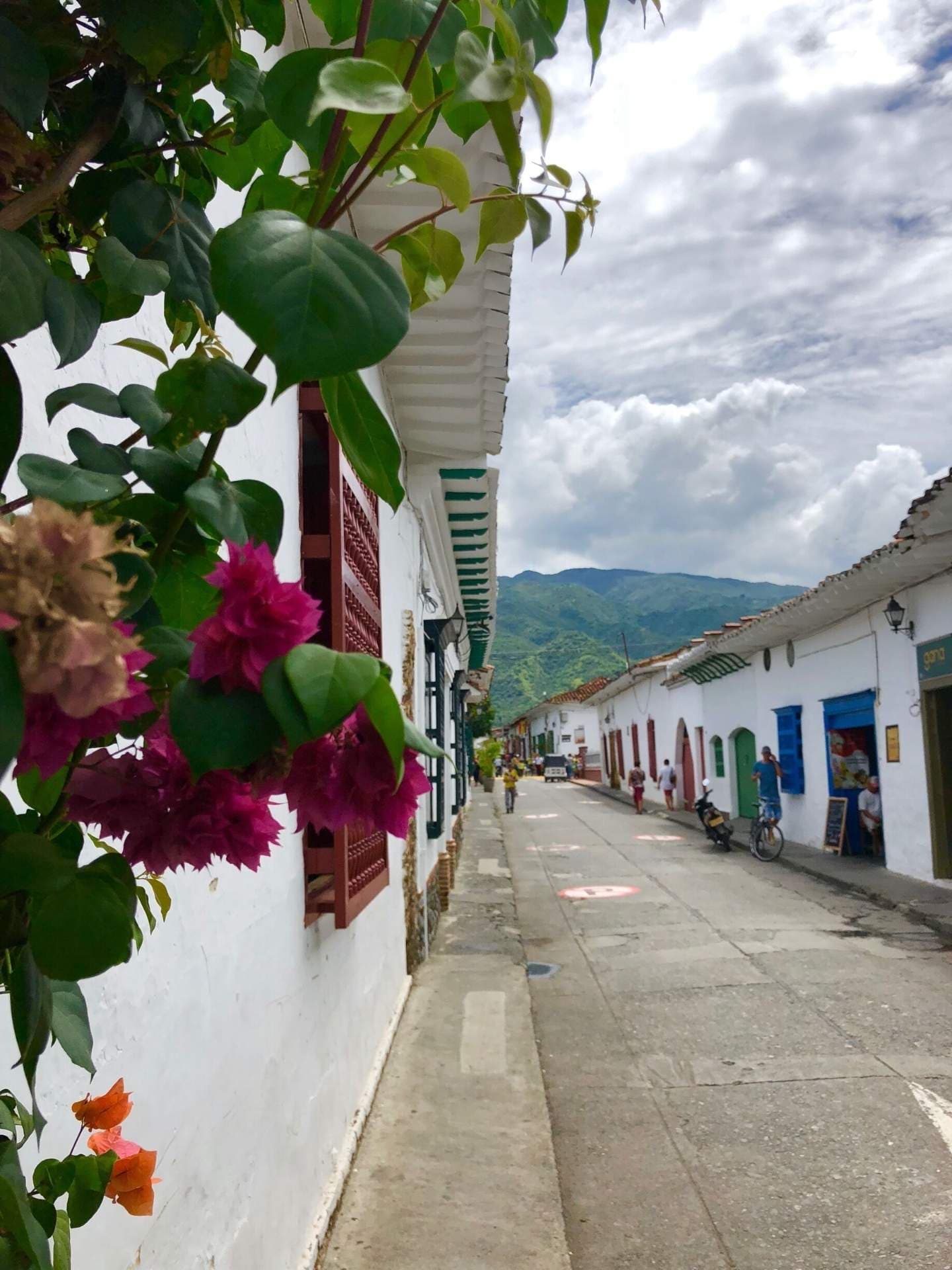 Street scene in Liborina with white buildings, colorful shutters, bougainvillea, distant mountains, and a few pedestrians.