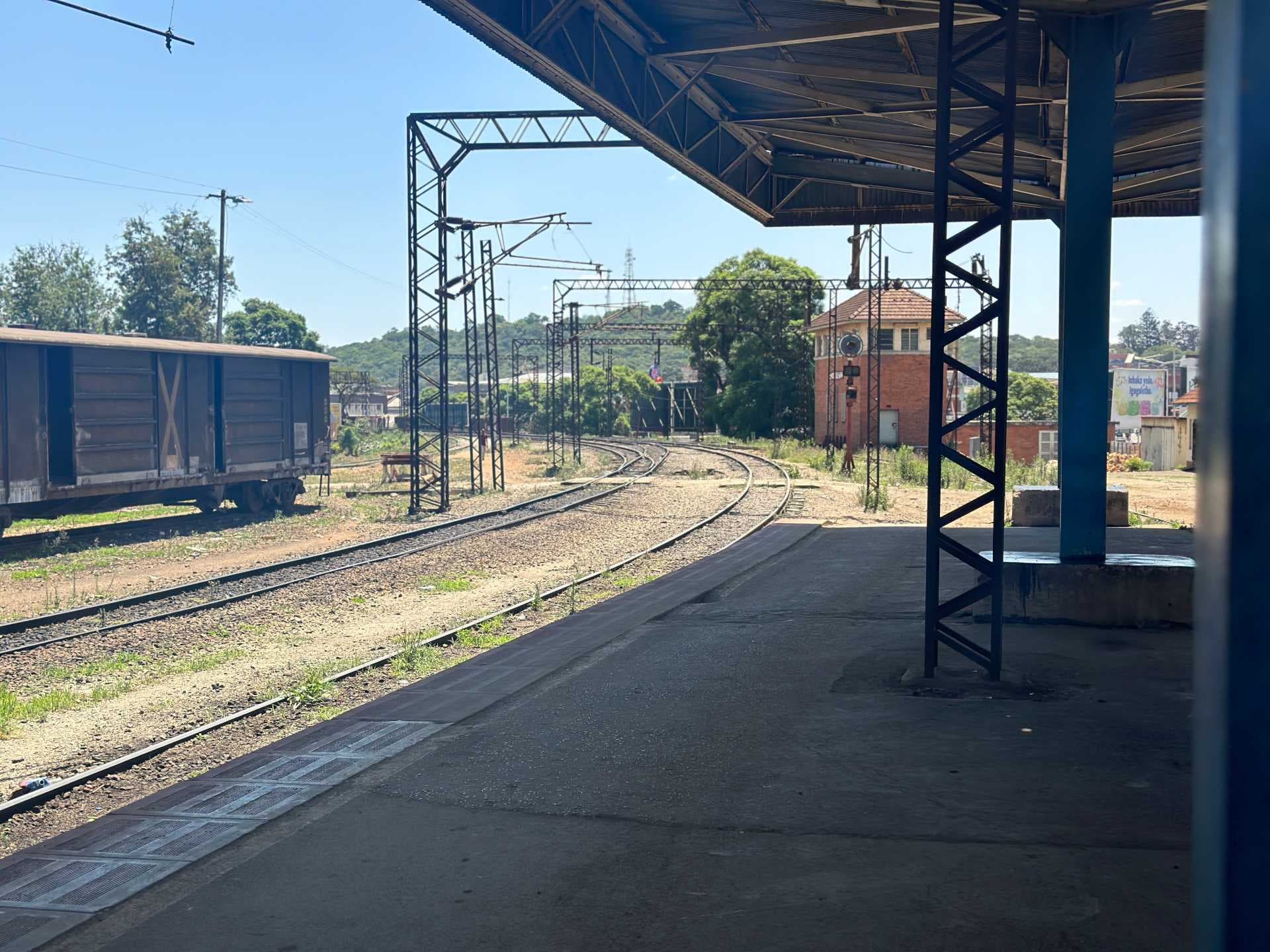 Railway station platform with curving tracks, overhead catenary structures, and a parked freight wagon under a clear sky.