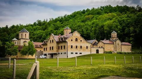 Wide exterior view of a large stone building complex with multiple towers, set against a green forested hillside near Kalenderovci Donji.