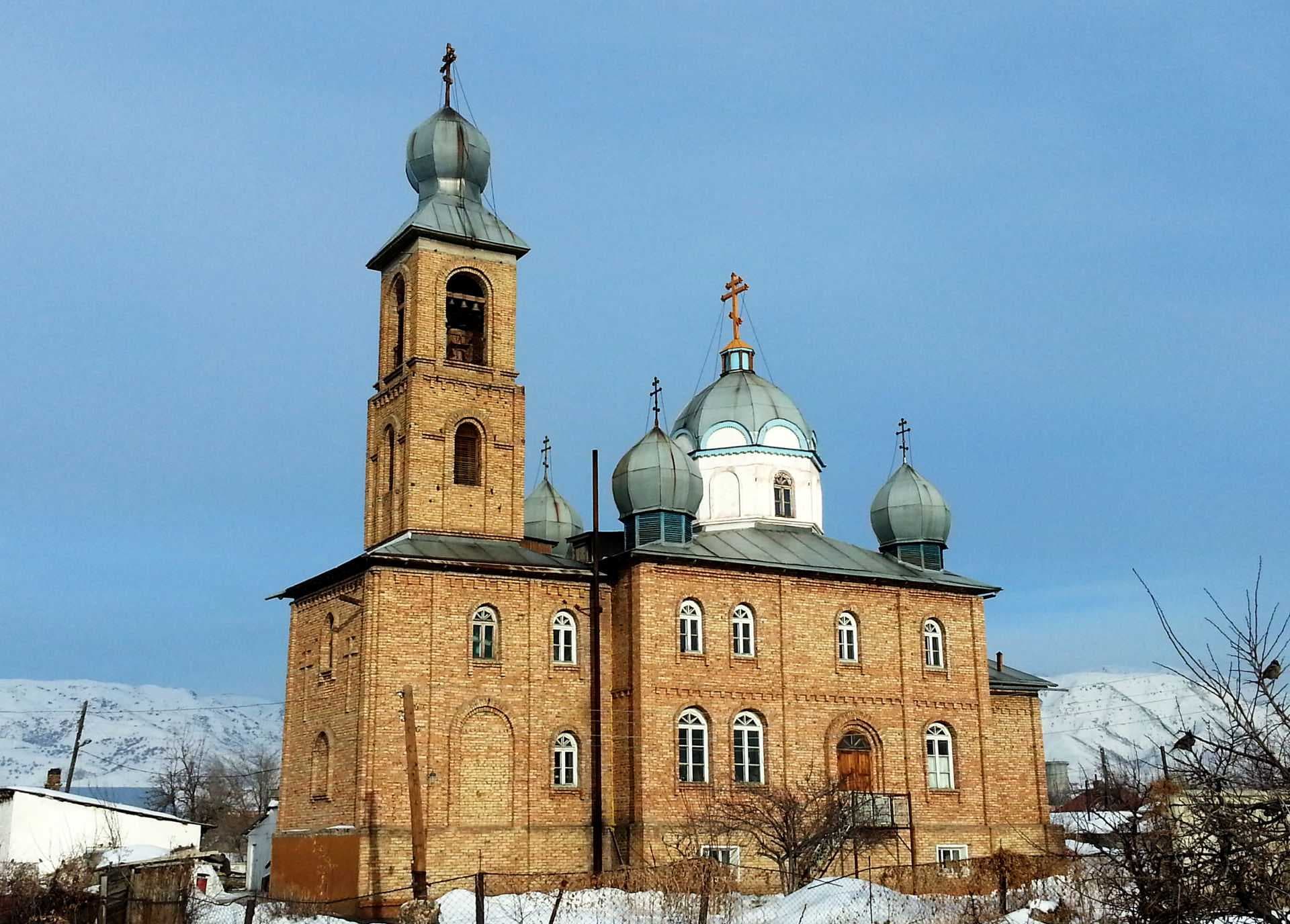 Brick Orthodox church with green domes and bell tower in a snowy small-town setting, mountains in background.