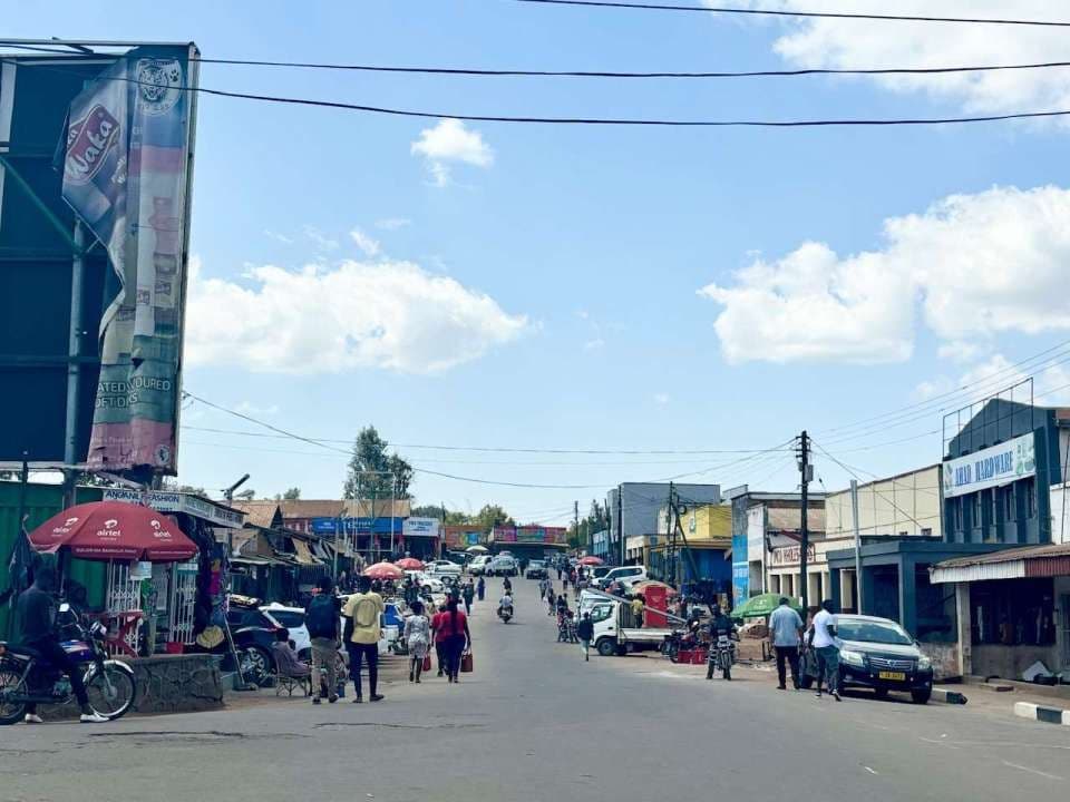 Wide street view in a town with shops, pedestrians, vehicles, and overhead wires under a partly cloudy sky.