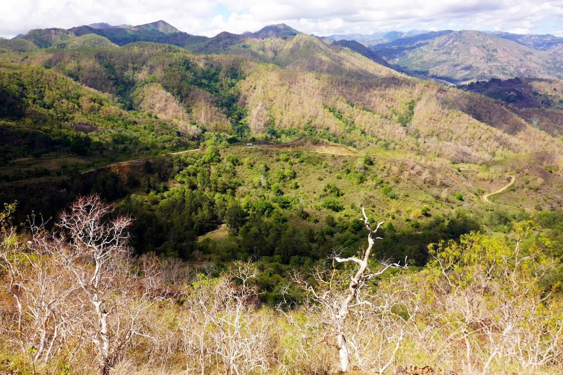 Wide landscape photograph of dry, hilly terrain with sparse vegetation, winding dirt roads, and distant mountains under a partly cloudy sky