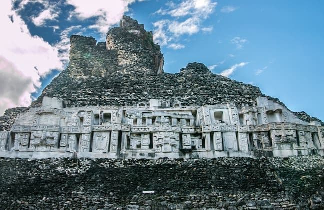 Maya stone temple structure with carved facade and stepped pyramid rising against a blue sky with scattered clouds.