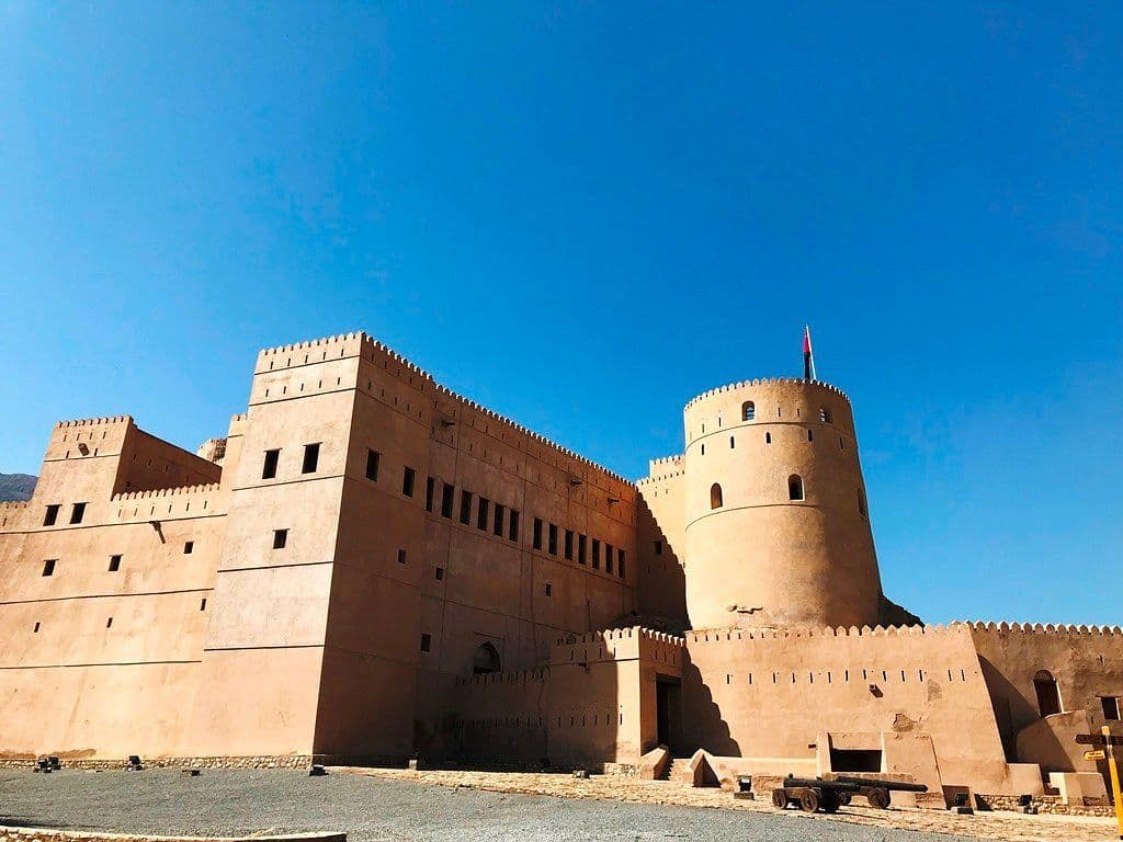 Wide exterior view of Rustaq Fort featuring massive sandstone walls, a prominent round tower, and a clear blue sky.