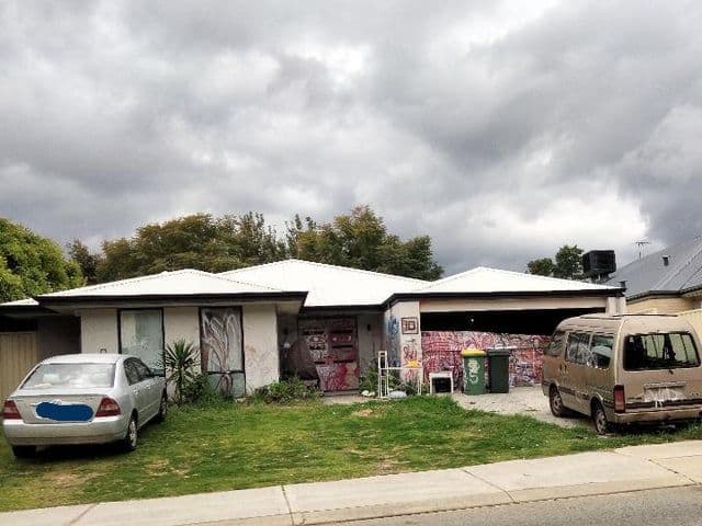 A silver sedan and a beige van are parked in the driveway of a suburban house with a white roof under an overcast sky in a Perth suburb.