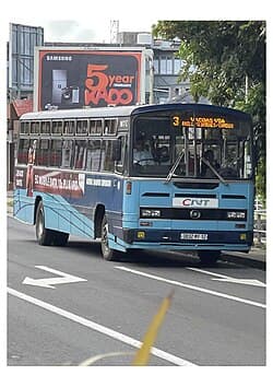A blue and white public bus with CNT branding drives on a paved road in Terre Rouge, Mauritius, passing a large billboard.