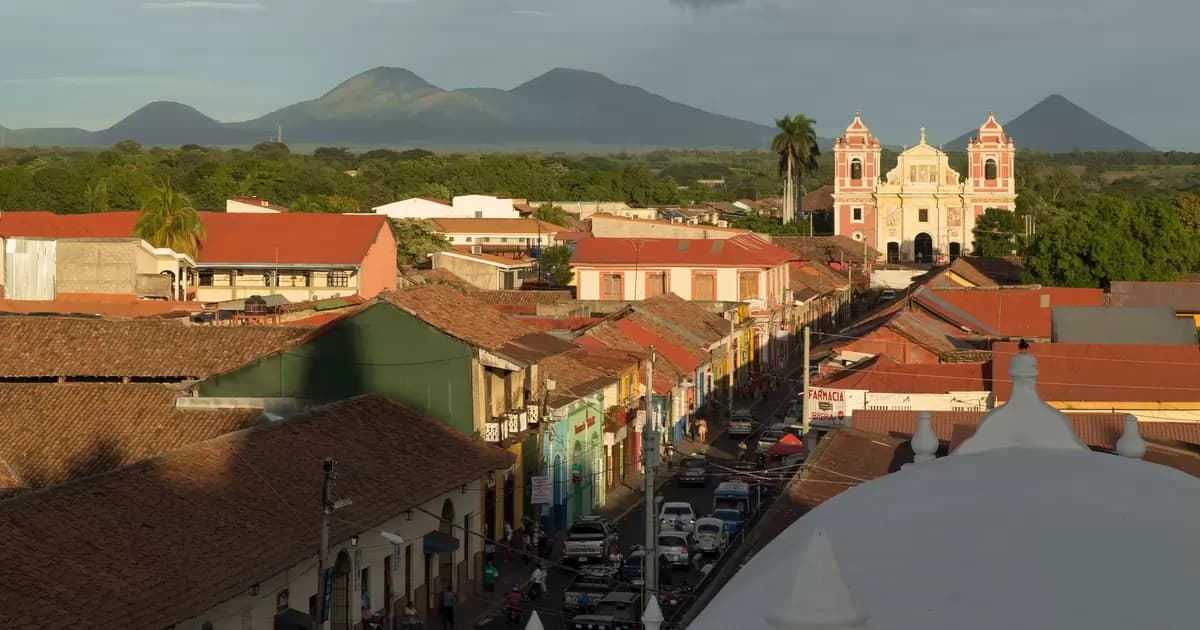 Wide elevated view of a colonial town with red-tiled roofs, a central street, and a prominent church, backed by distant mountains.