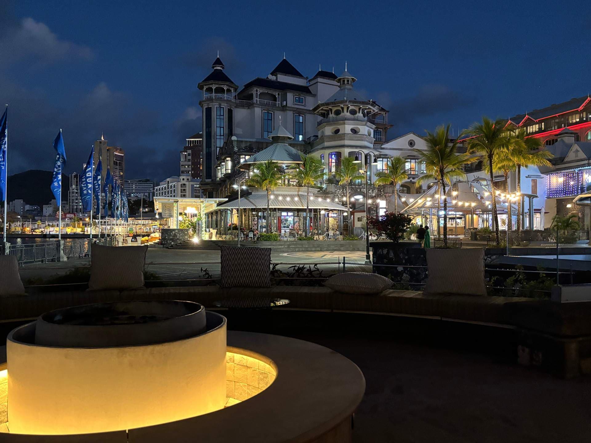 Nighttime waterfront plaza in Port Louis with illuminated colonial-style buildings, palm trees, flags, and outdoor seating.