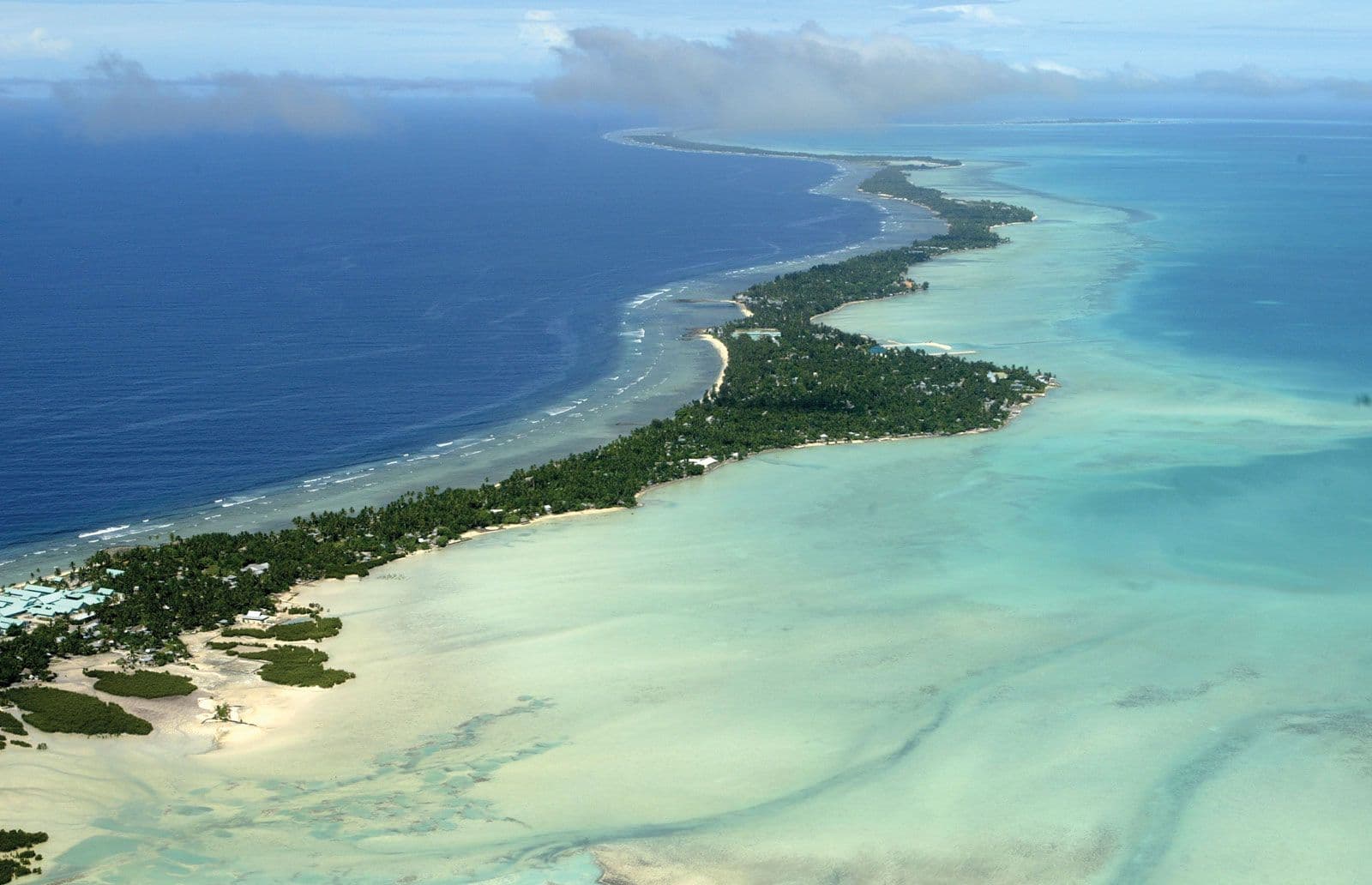 Aerial view of a long narrow tropical atoll island fringed by turquoise lagoon and deep blue ocean with palm groves and scattered buildings along the coastline