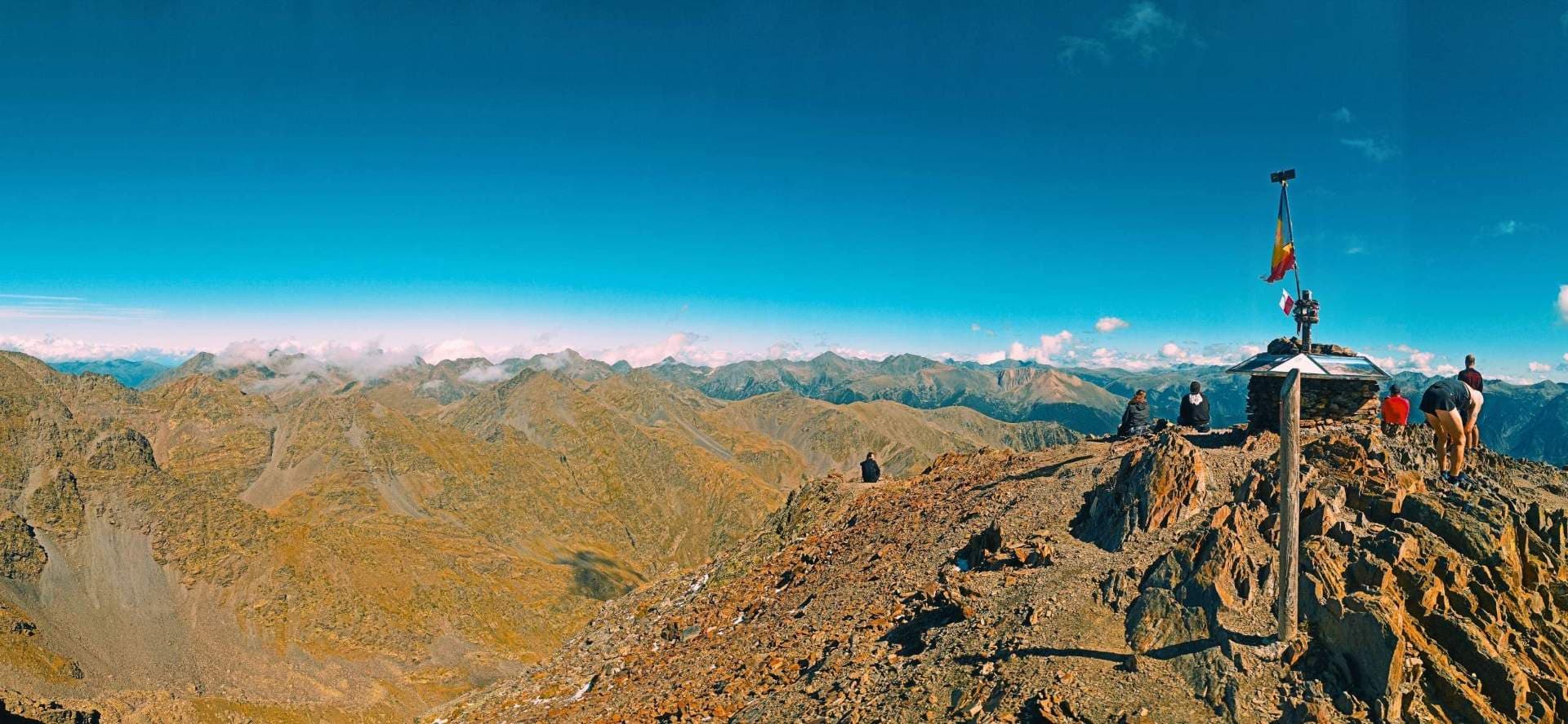 Wide mountain summit panorama with hikers near a stone marker and flags, overlooking rugged ridges under a clear blue sky.