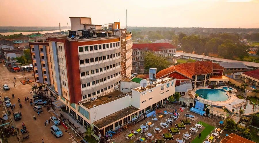 Aerial view of a multi-story hotel complex with a swimming pool and outdoor seating area, situated near a river and surrounded by local streets.