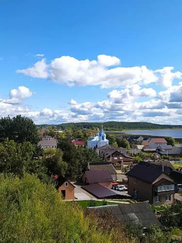 Elevated view of a small Belarusian town with a blue-domed church, residential houses, and a lake under a partly cloudy sky.