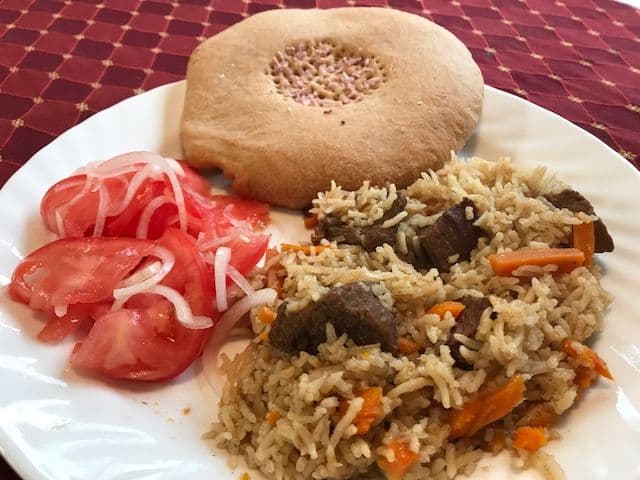 A plate of Tajik plov with rice, carrots, and meat, served with tomato-onion salad and a round flatbread on a patterned tablecloth.