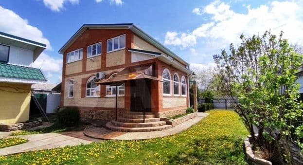 Exterior view of a two-story brick and stucco house with a green roof, set in a grassy yard with dandelions under a blue sky in Krasnodar Territory.