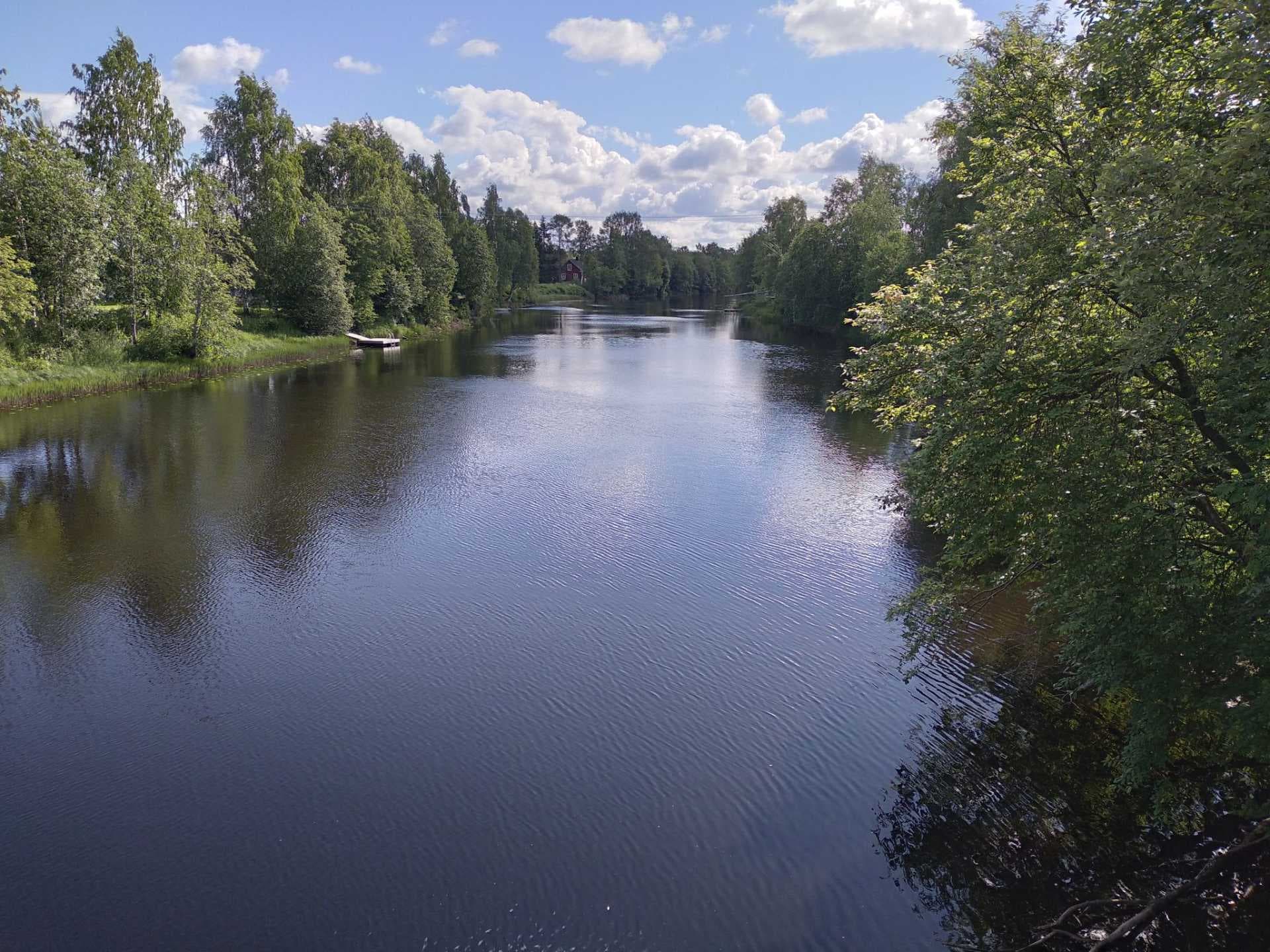A serene river landscape in the Kaustinen region, featuring calm waters reflecting a blue sky, flanked by lush green trees and a distant red house.