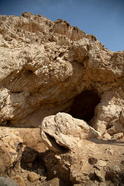 Sunlit rocky cliff face featuring a dark cave entrance and scattered boulders, depicting ancient archaeological ruins at Mleiha.